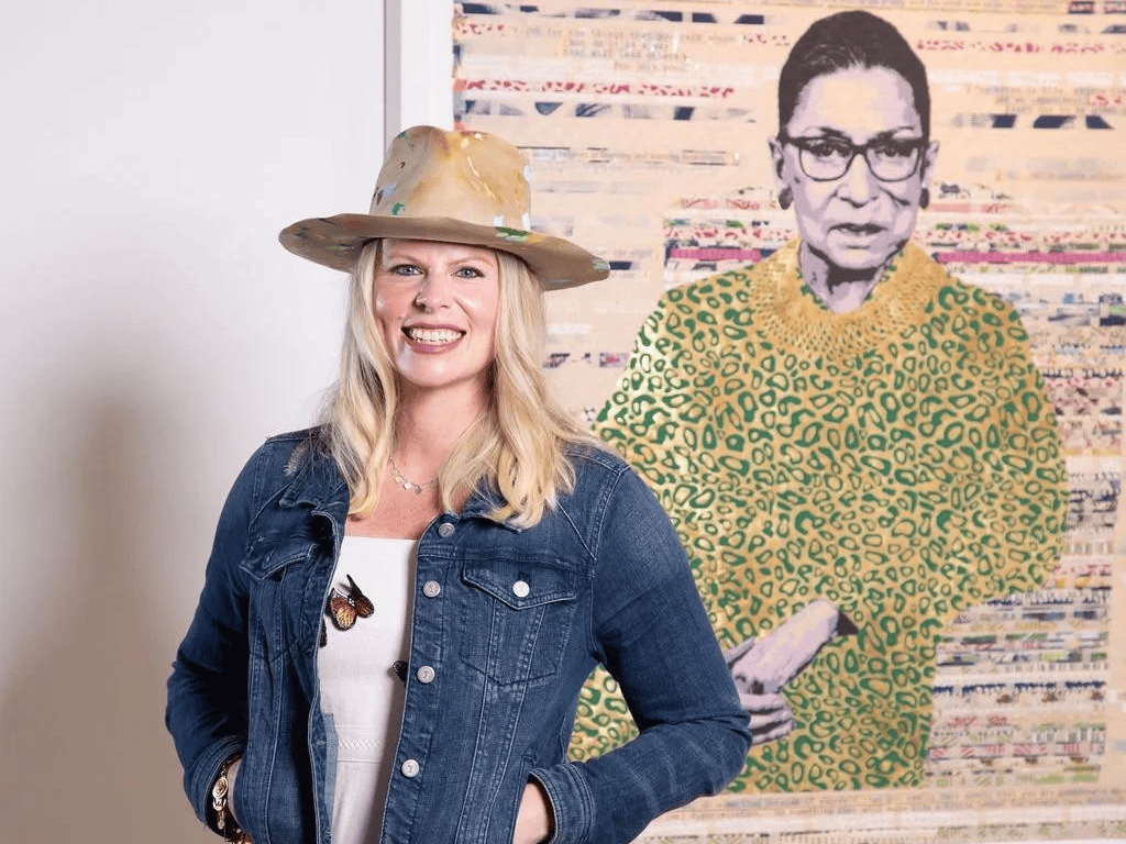 Woman in a denim jacket and hat standing beside a pop-art portrait in a gallery