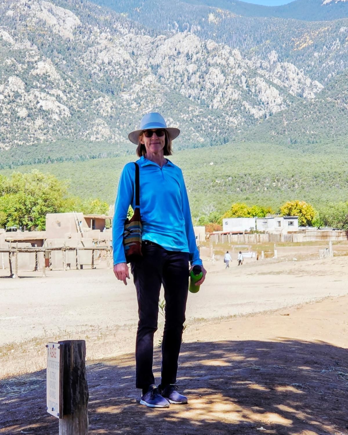 Person in blue shirt and sun hat standing outdoors with mountains and buildings in the background.