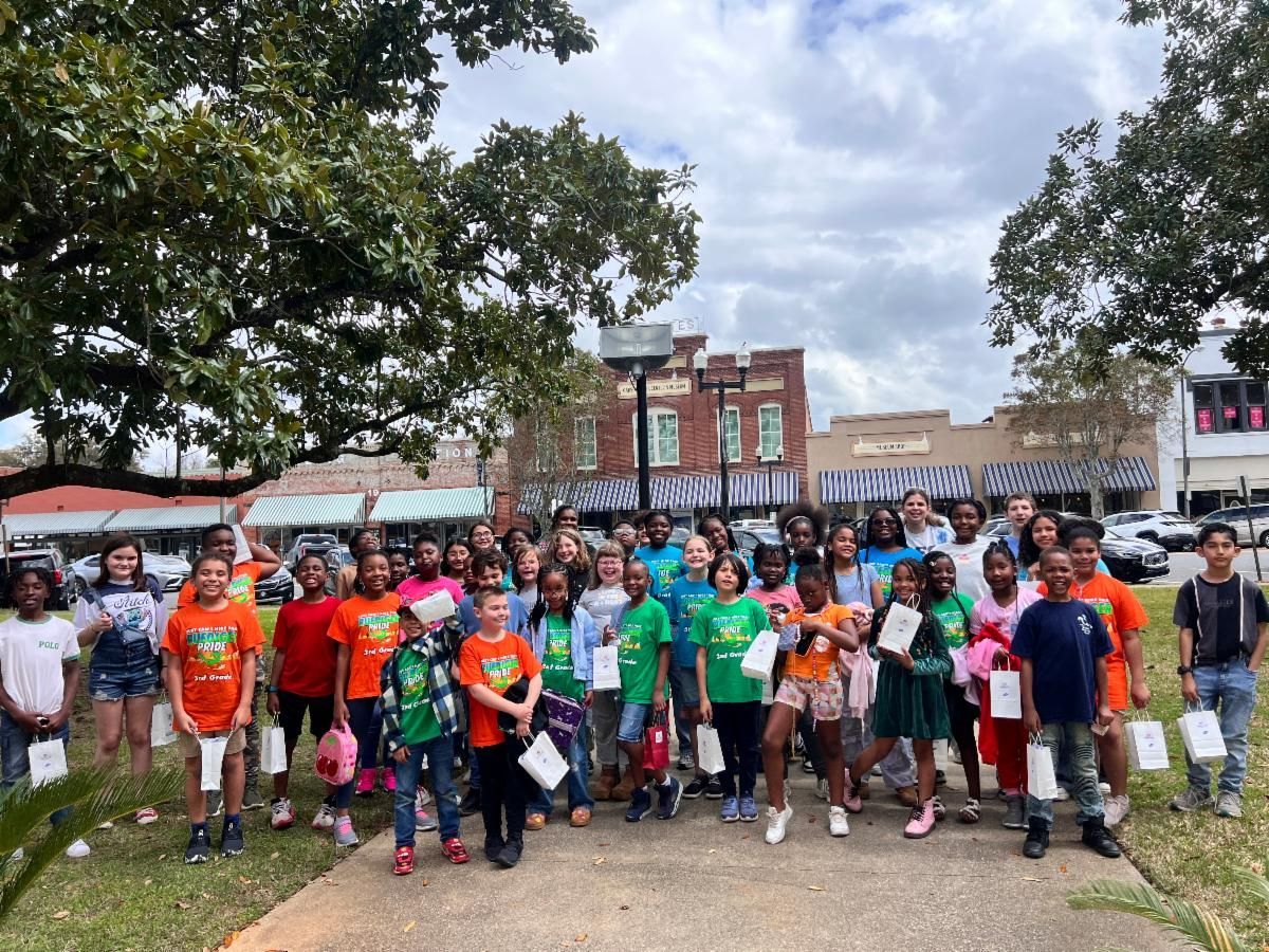 Group of children and adults posing outdoors in front of buildings, many wearing bright orange and green shirts.