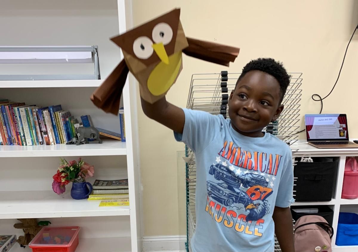 Child holding up a homemade owl craft in a classroom, smiling beside shelves and cubbies.