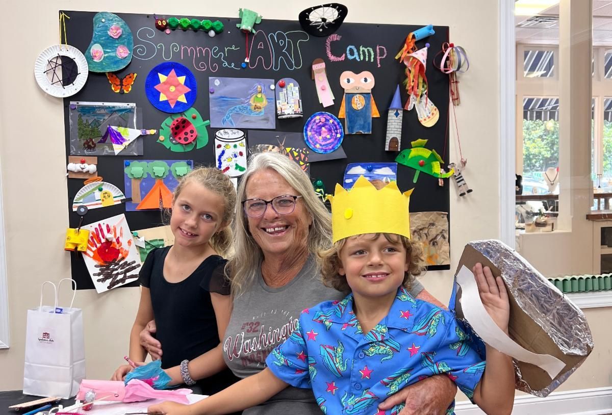 Woman and two children smiling at a craft table with colorful artwork and paper hats displayed behind them