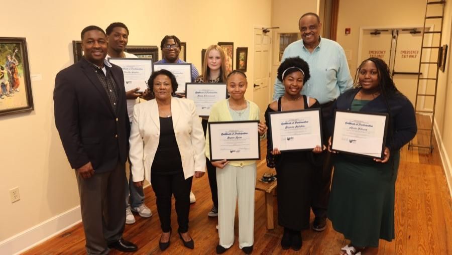 Group of people posing indoors with framed certificates in a hallway, smiling for a recognition photo