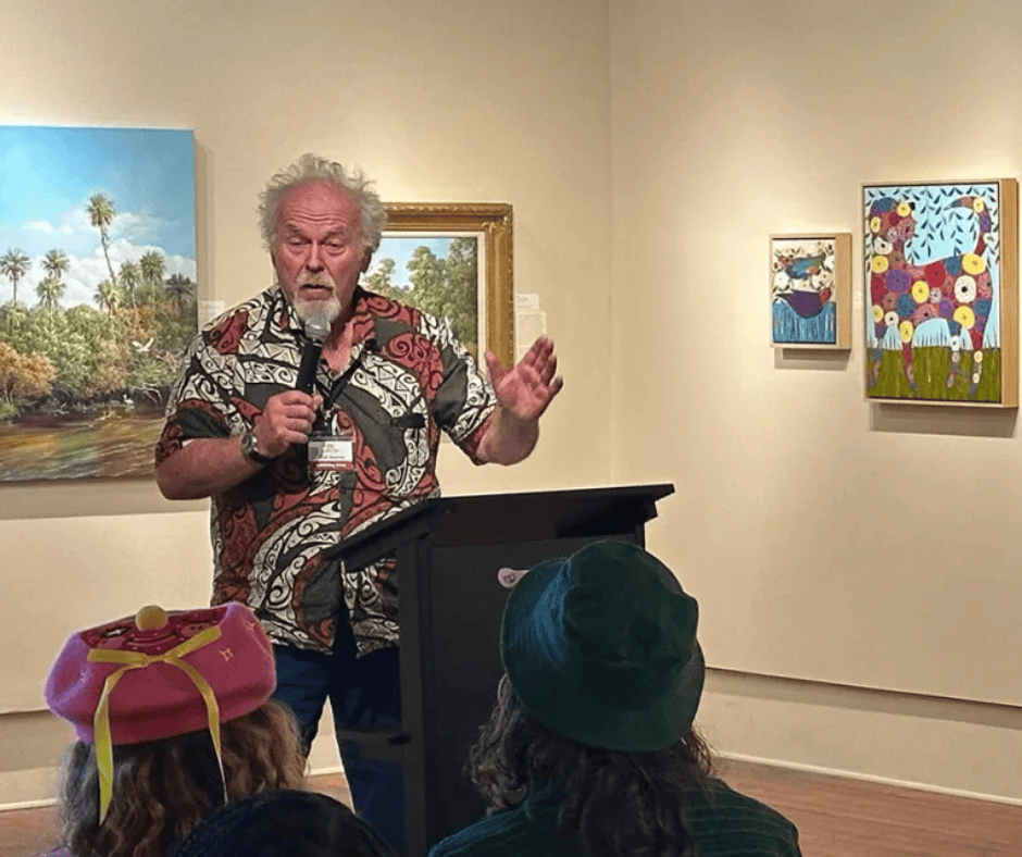 Speaker at a podium in an art gallery, gesturing in front of colorful paintings.