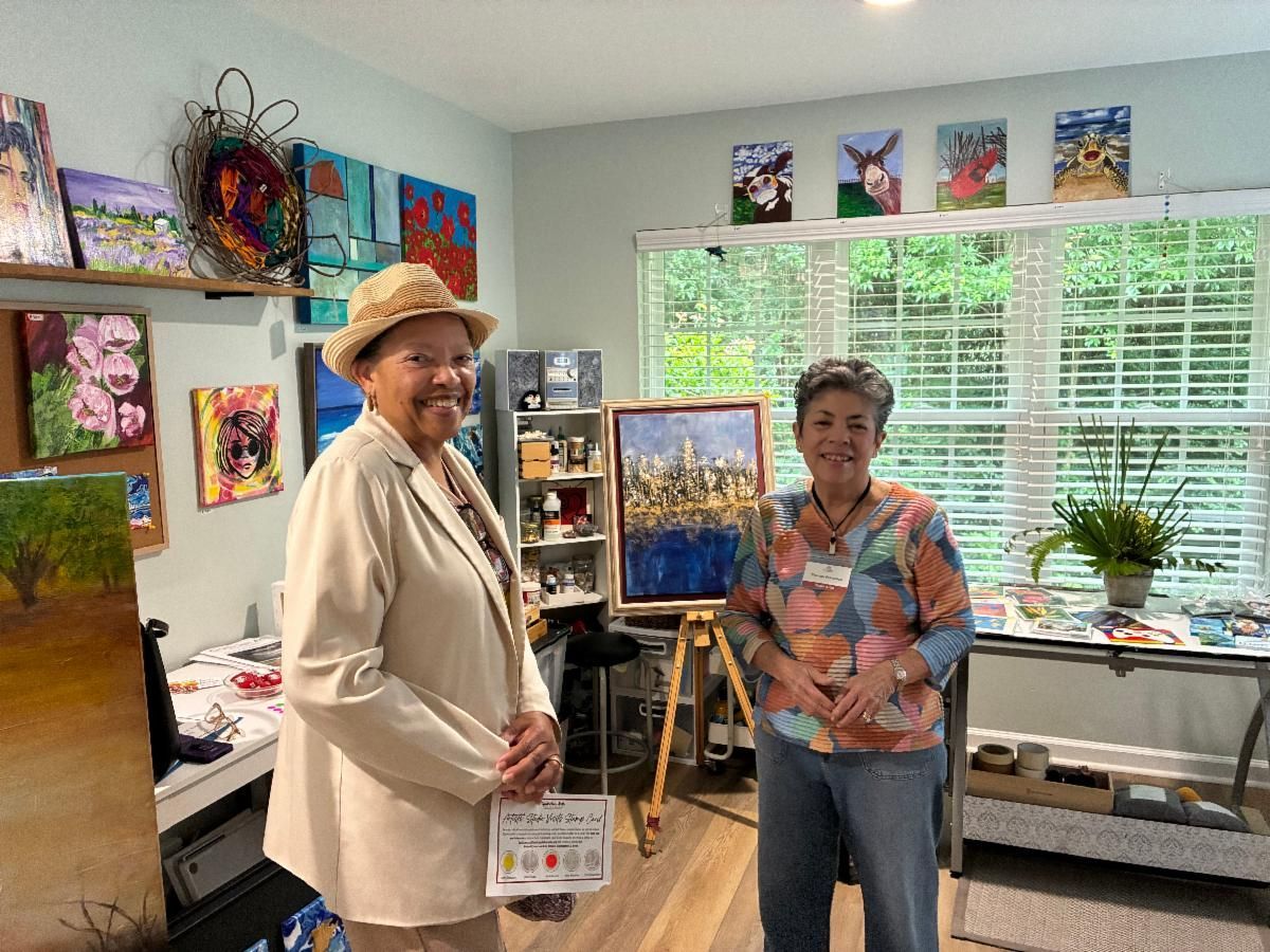 Two people smiling in an art studio, holding a painting near colorful artworks and a bright window.