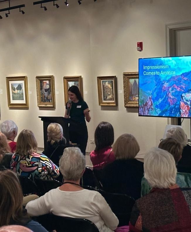 Audience watching a woman speak in an art gallery, with framed paintings and a presentation screen beside her