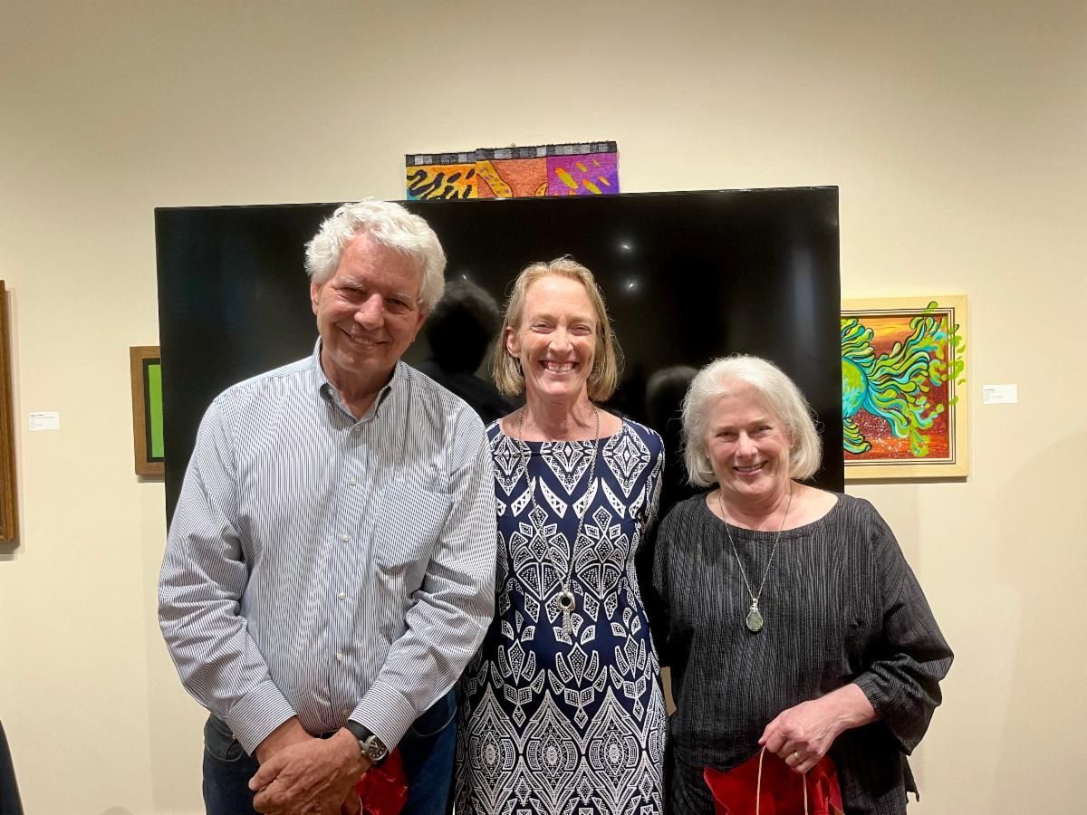 Three people smiling indoors in front of a large black display and colorful artwork.