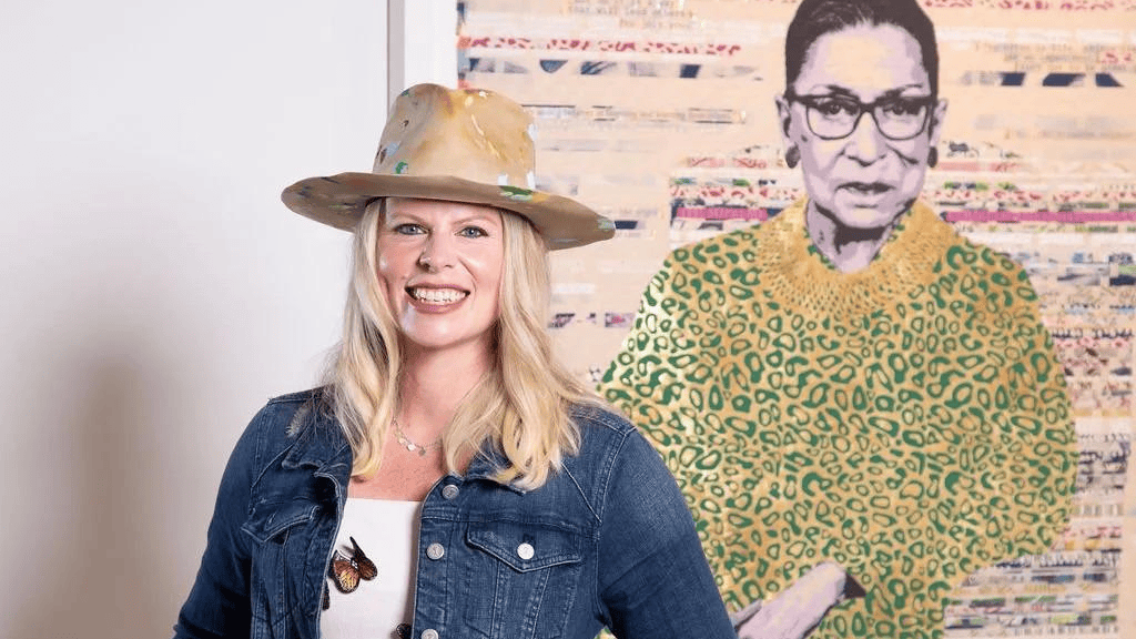 Smiling woman in a tan hat and denim jacket stands beside a colorful portrait mural.