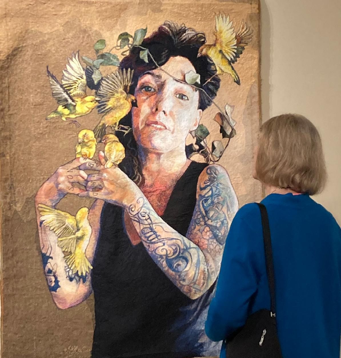 Art gallery painting of a tattooed figure with birds on a canvas, viewed beside an older visitor in blue.