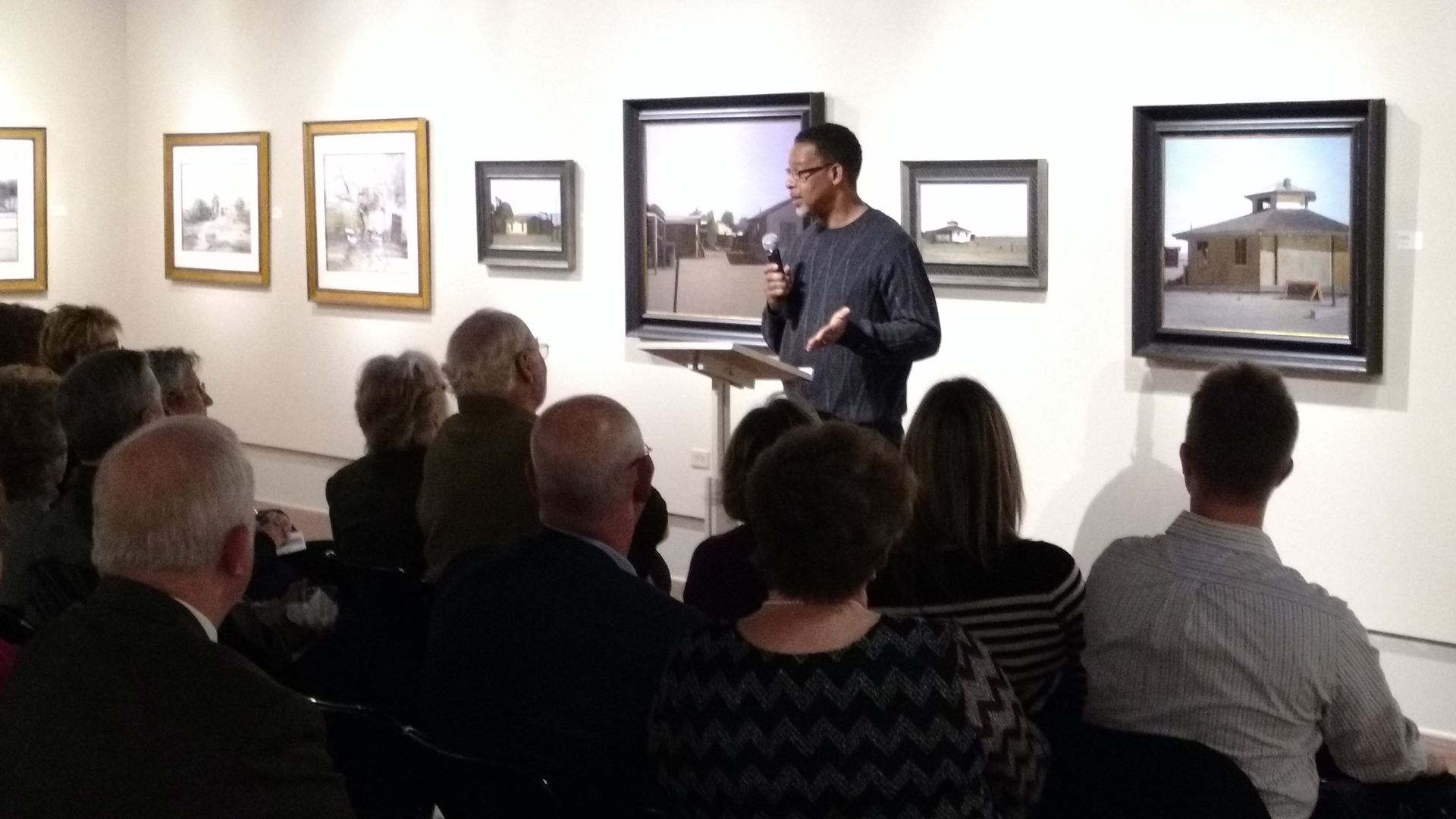 Gallery talk with an audience seated before framed artwork, as a man gestures beside a projected image.