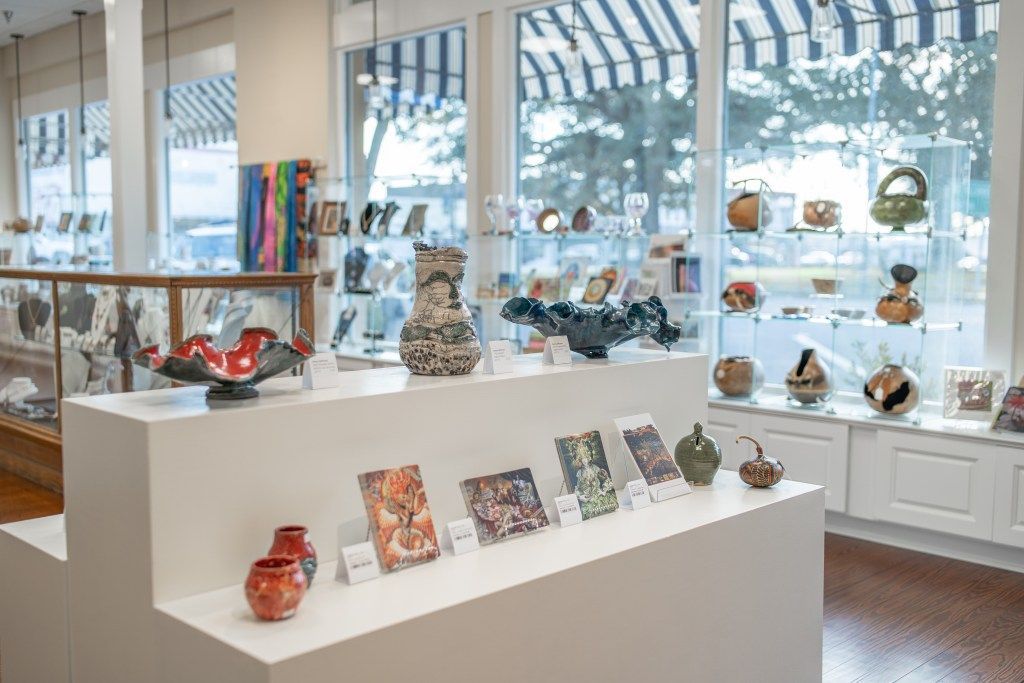 Bright gallery display of colorful glass art on white pedestals before large windows