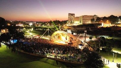 Crowd gathered around a lit stage at dusk in a park, with city buildings glowing in the background.