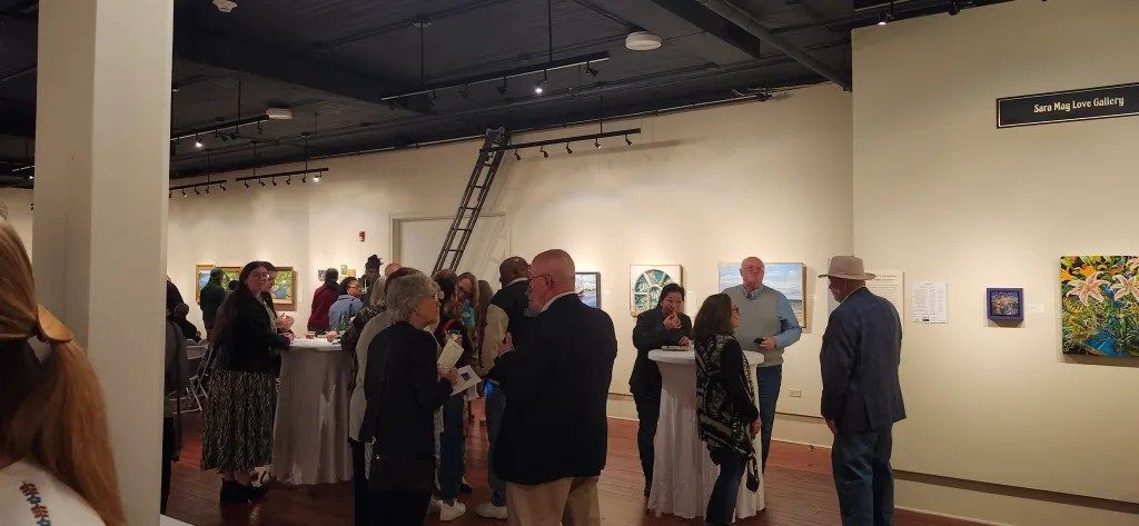 People mingling in an art gallery with paintings on white walls and a ladder overhead