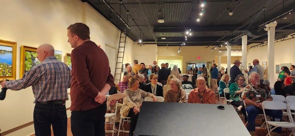 Indoor gathering with people seated at tables and two speakers near a wall display, viewed from the front stage