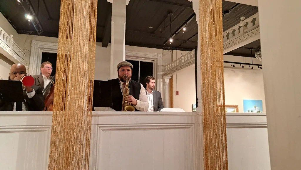 Three people stand behind a white display counter in an indoor gallery with hanging beige fabric panels.