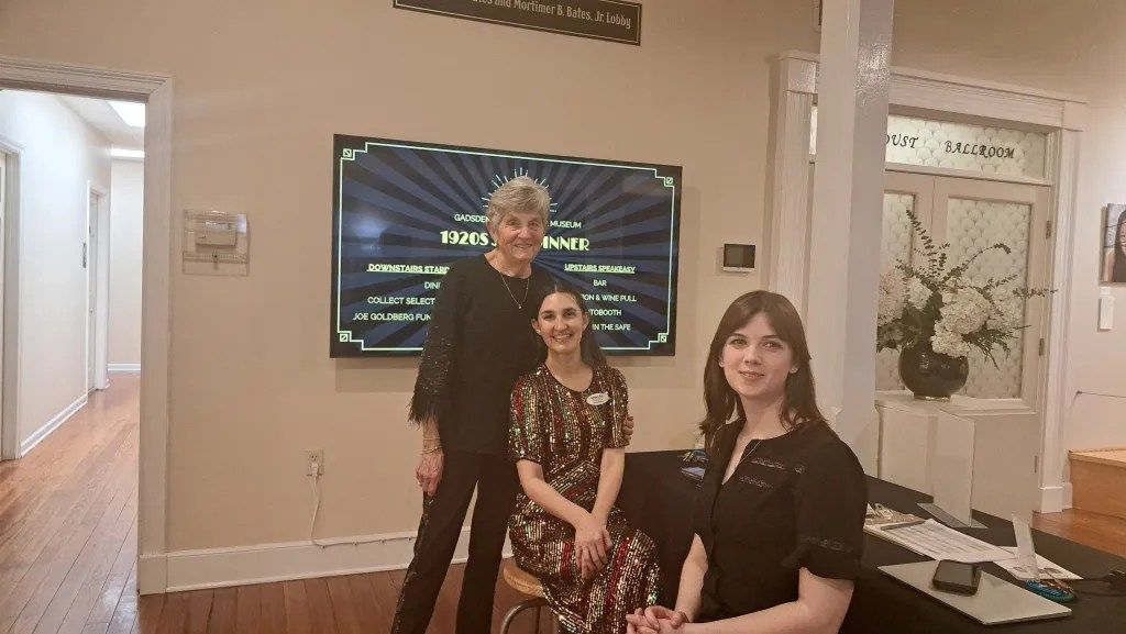 Three people pose in a room beside a large screen and keyboard, with floral decor in the background.