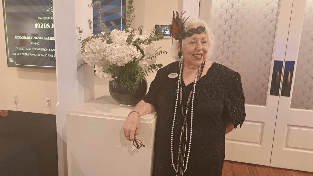 Woman in black dress standing indoors beside white flowers and a doorway, smiling at an event.