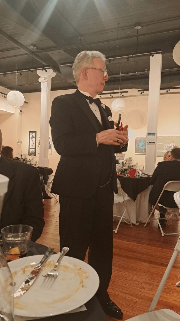 Man in a dark suit speaking at a banquet table in a decorated event hall