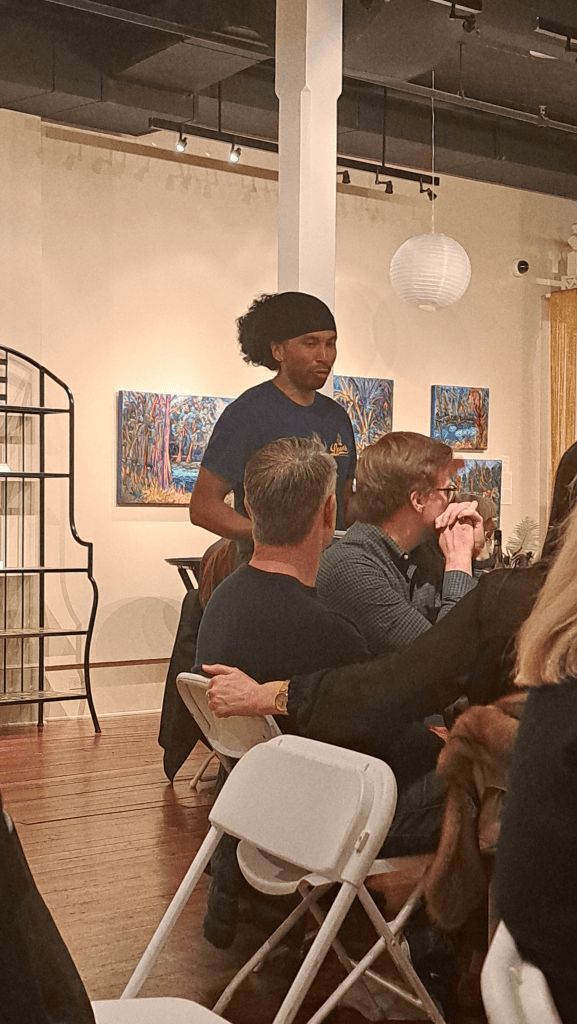 People seated in a gallery while a speaker stands near colorful wall art and a hanging white sphere.