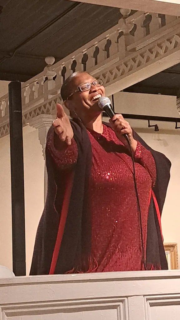 Woman in a red outfit speaking into a microphone on a balcony, gesturing with her free hand.
