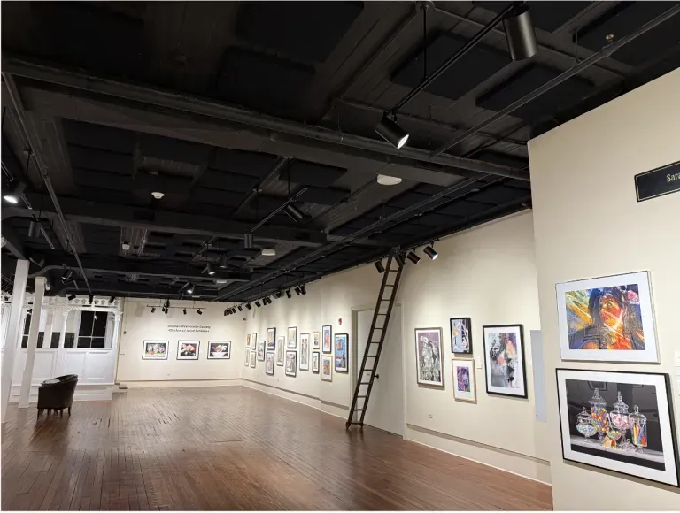 Art gallery interior with framed paintings on white walls, wooden floor, and a ladder under a black ceiling