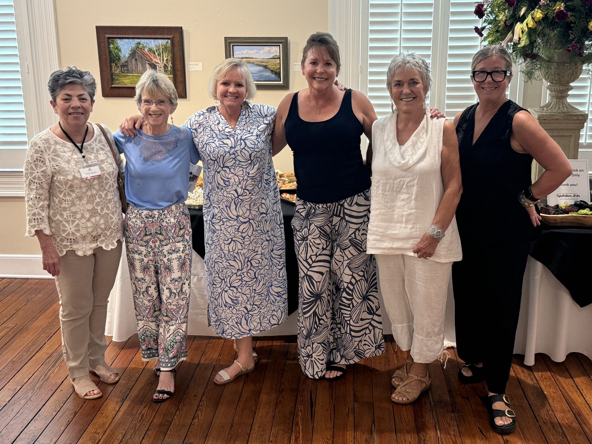 Six women posing together in a decorated indoor room, dressed in light and black outfits.