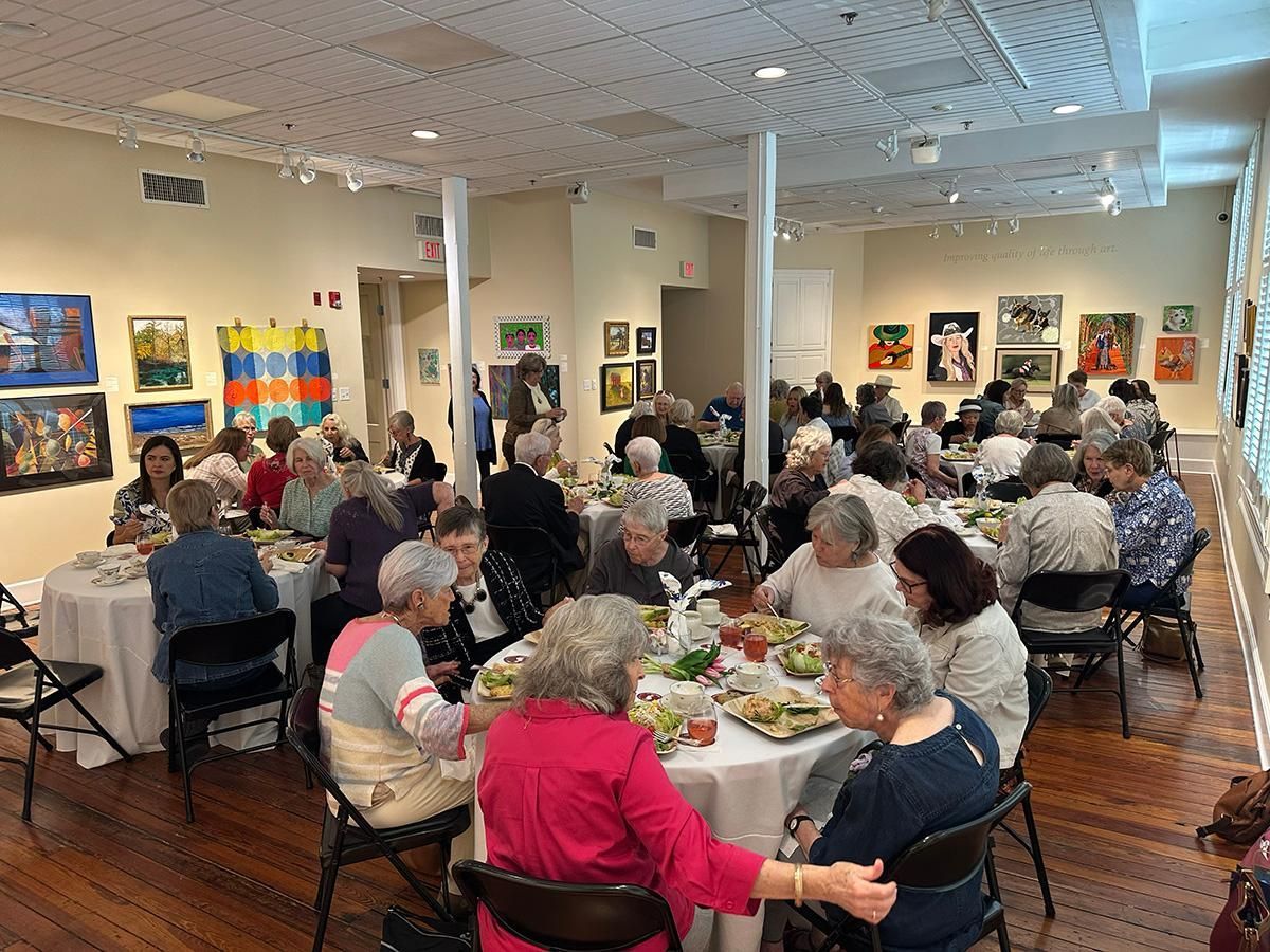 Packed art gallery gathering with seated guests at tables in a bright room with framed paintings on the walls