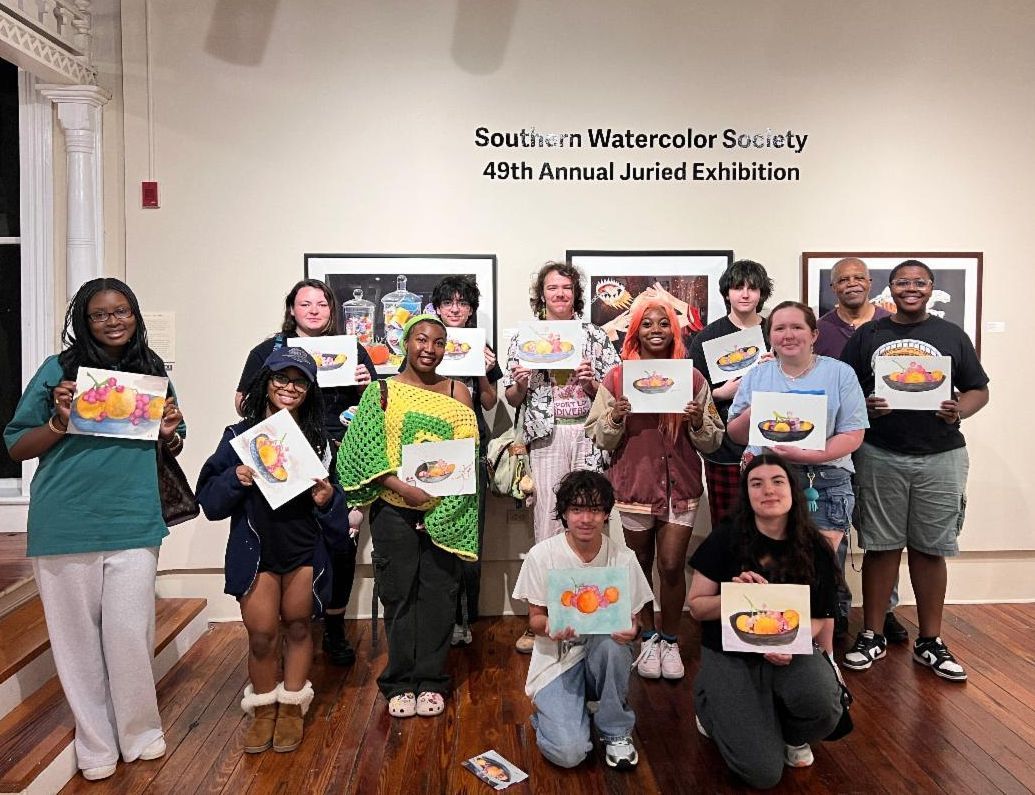 Group of people holding colorful paintings in a gallery-like room, posing in two rows.