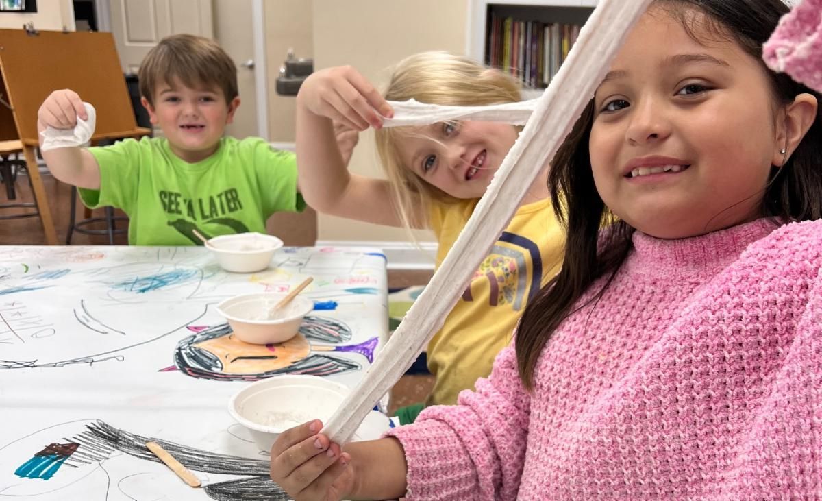 Children smiling and painting at a table, with brushes, paper, and art supplies in a classroom.