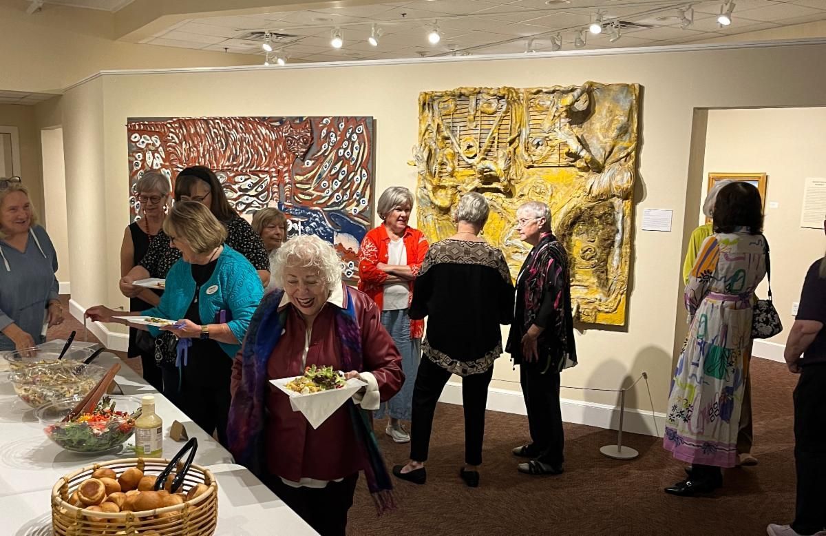 Art gallery reception with people mingling near buffet tables and colorful abstract paintings on the walls
