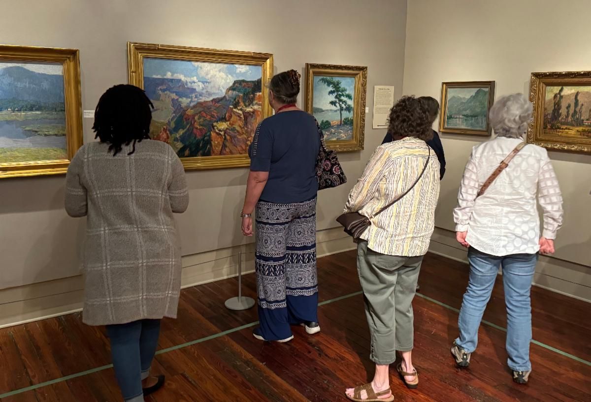 Four people view framed landscape paintings in a gallery with wooden floors.