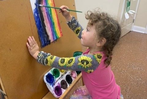 Child painting a rainbow on a hanging paper strip with a brush, using watercolor paint pots nearby