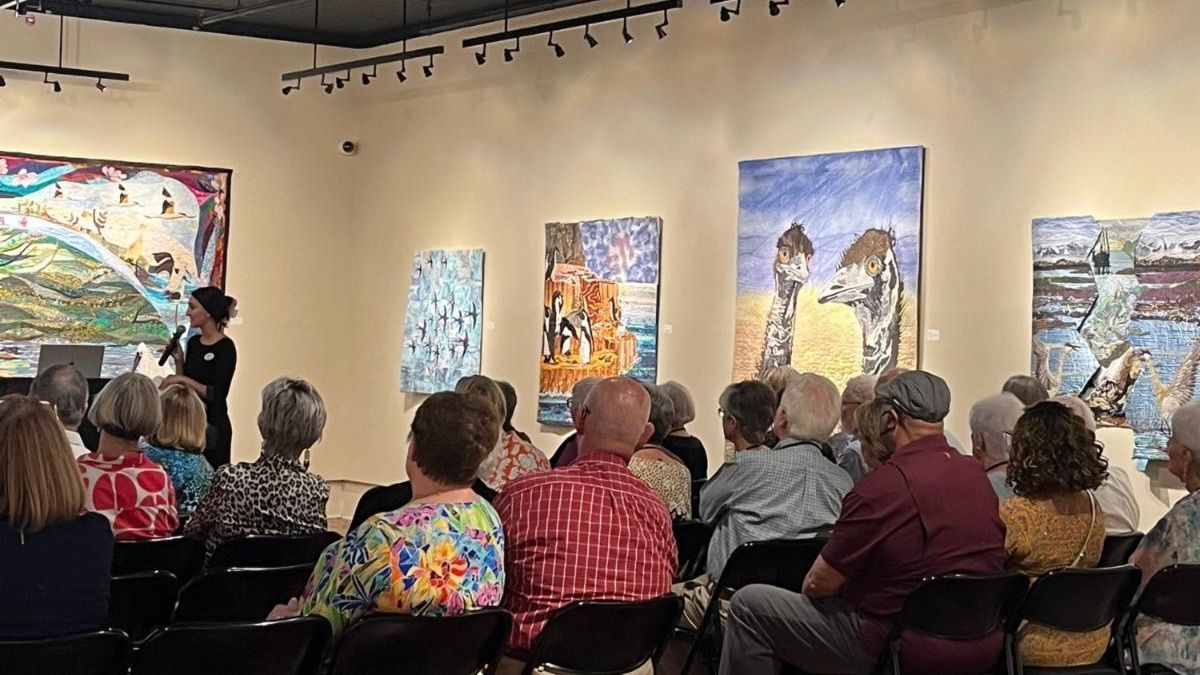 Audience seated in gallery listening to a presenter beside colorful paintings on a wall