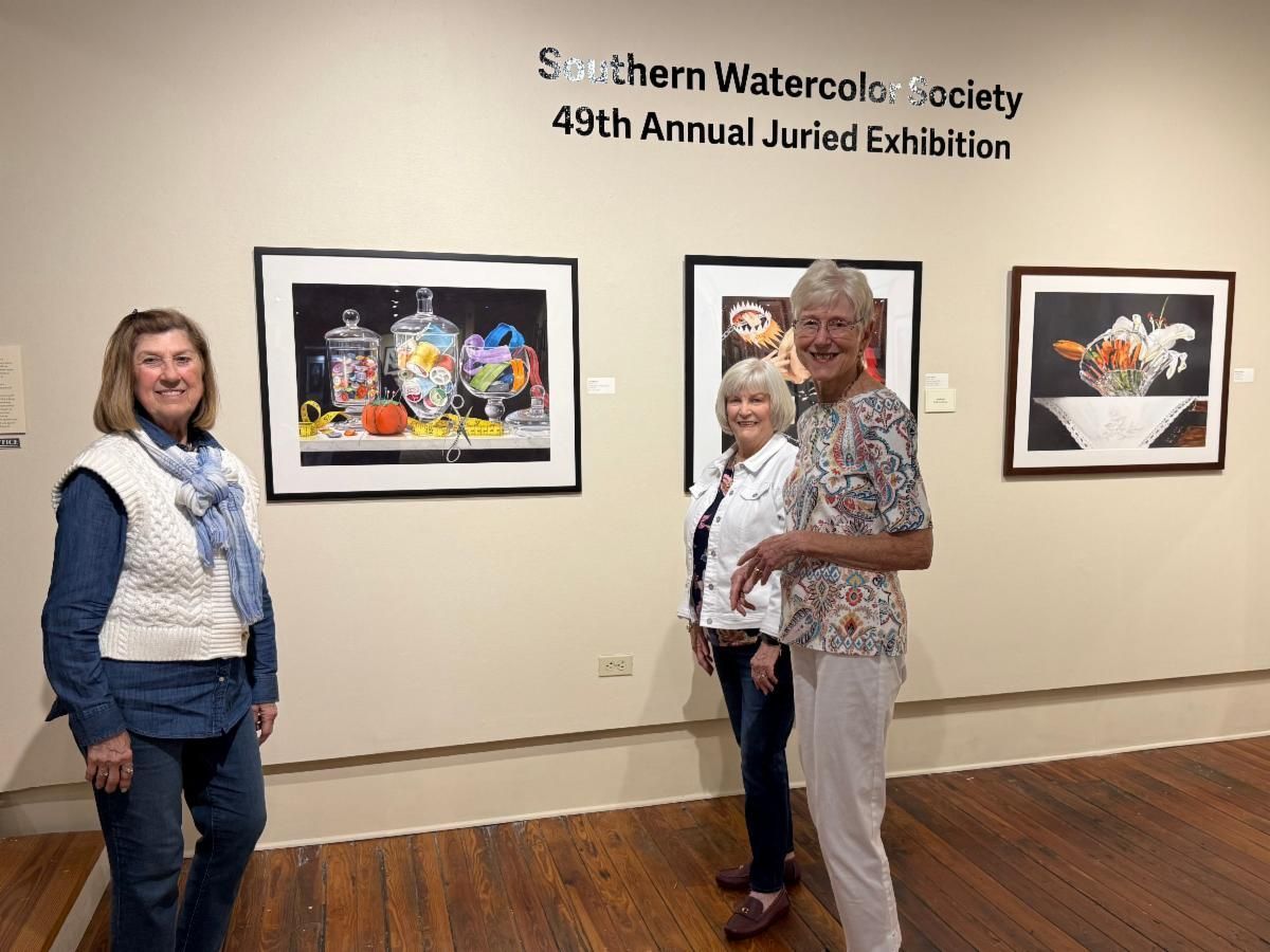 Three women standing in an art gallery beside framed watercolor paintings and an exhibition sign.