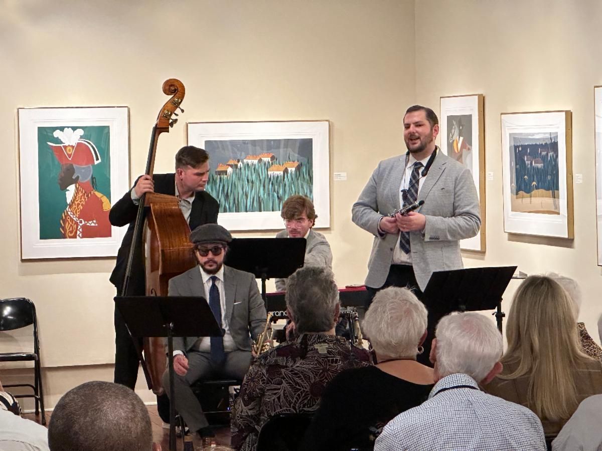 Small jazz ensemble performing in an art gallery before seated audience, with framed paintings on the walls