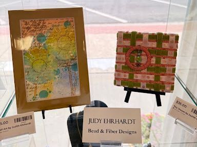 Display of colorful framed artwork and beaded textile pieces on easels at a craft show