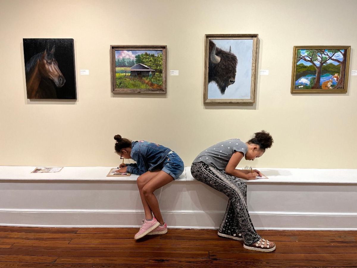 Two people sit back-to-back on a bench beneath framed paintings in a gallery.