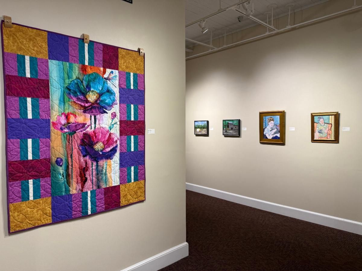 Gallery hallway with colorful quilt artwork and smaller framed paintings on beige walls.
