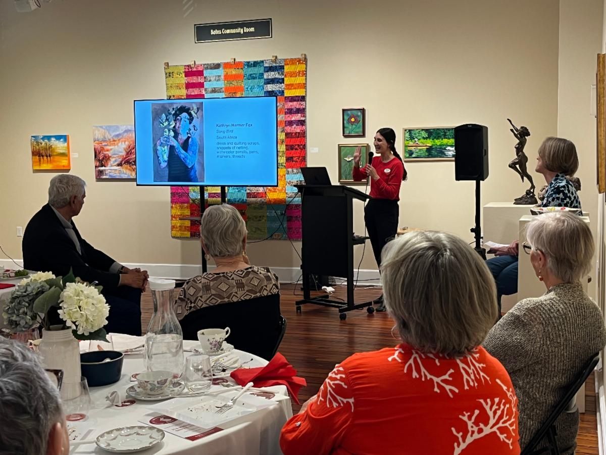 Presenter speaks beside a large screen at a community gathering with seated attendees at round tables.