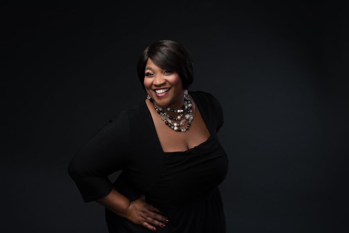 Smiling woman in a black dress and statement necklace against a dark background