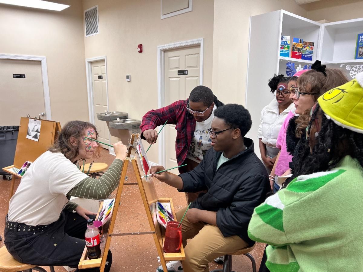 People seated at a long table in a classroom, interacting across a divider with art supplies visible.