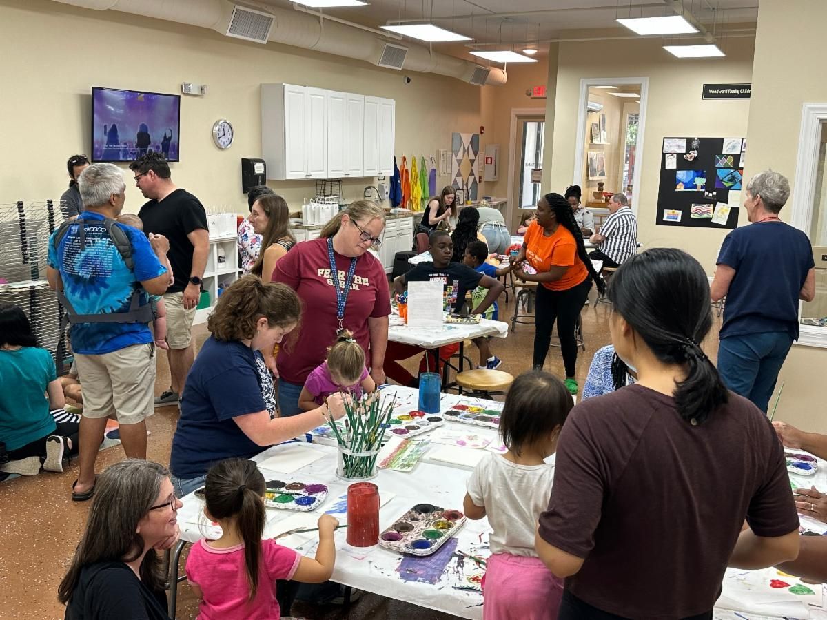 Busy community room with people gathered around tables at a craft activity, with supplies and wall art visible.