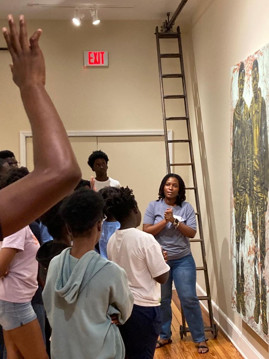 People gathered in an art gallery listening to a speaker beside a large wall painting and ladder.