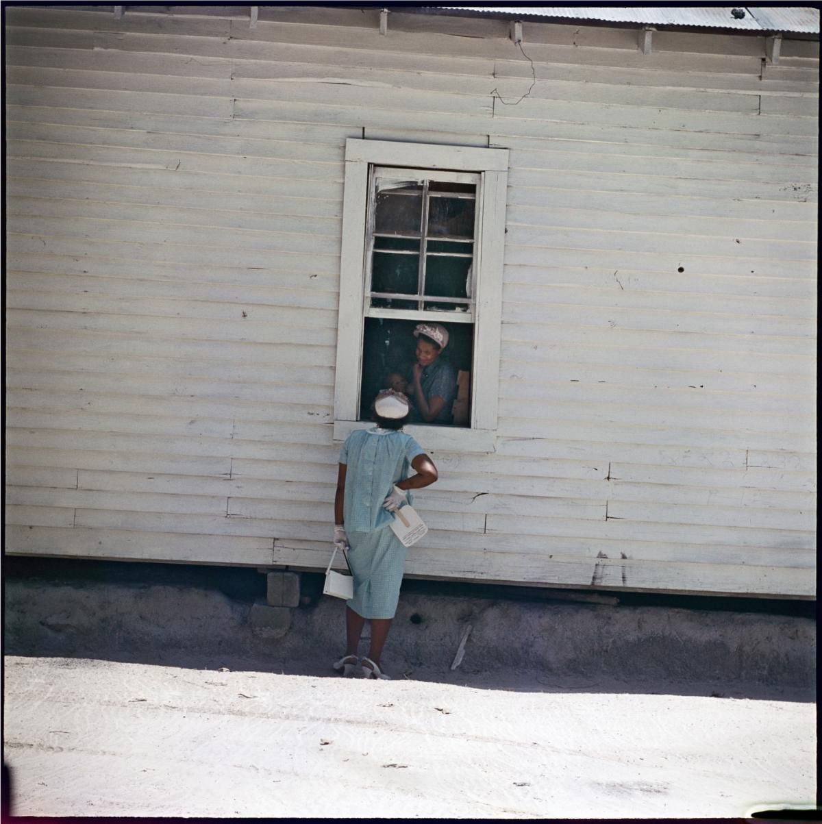 Person standing outside a white wooden house, looking up at someone in the window.