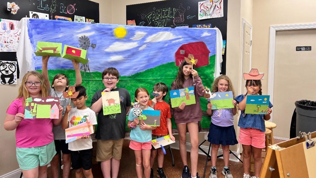 Children holding colorful art projects stand in front of a painted classroom mural.