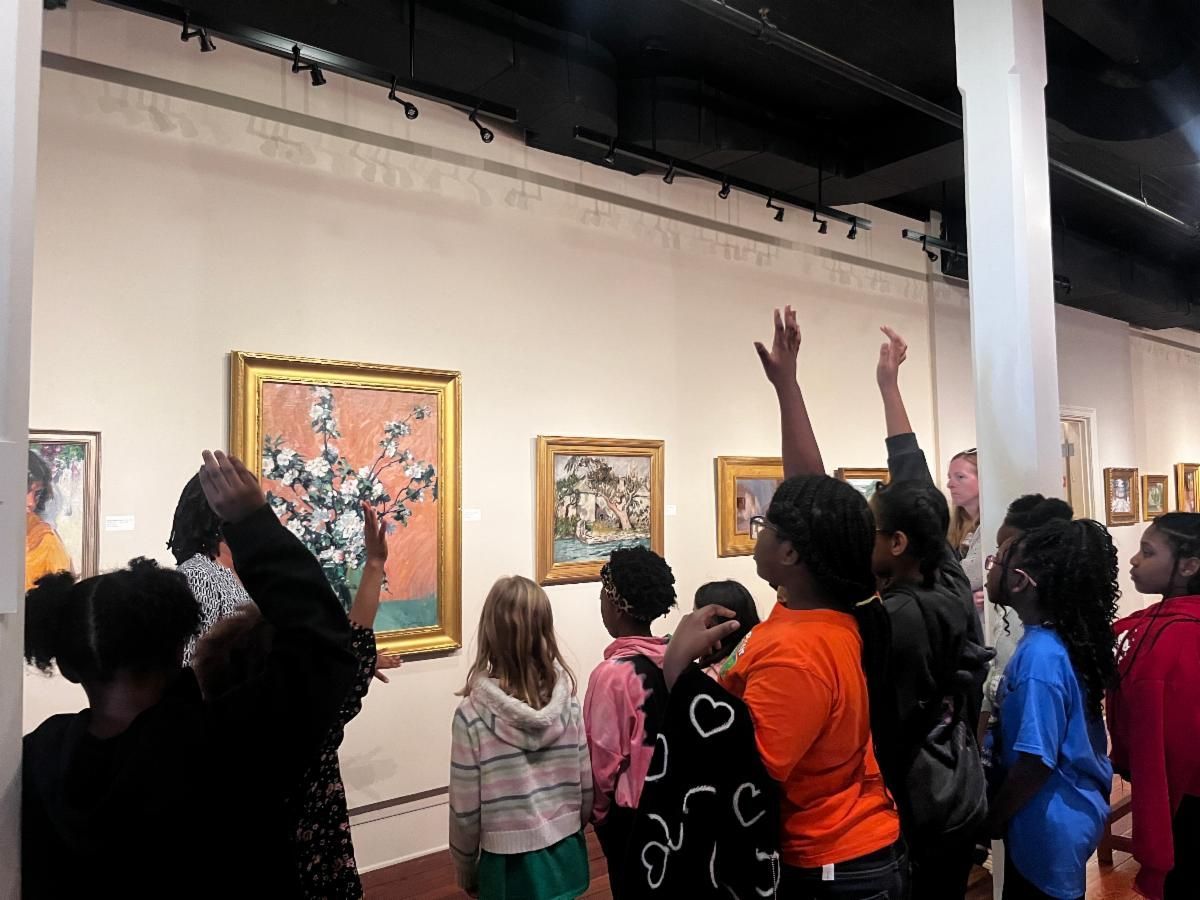 Visitors raising hands in an art gallery, viewing framed paintings on white walls.