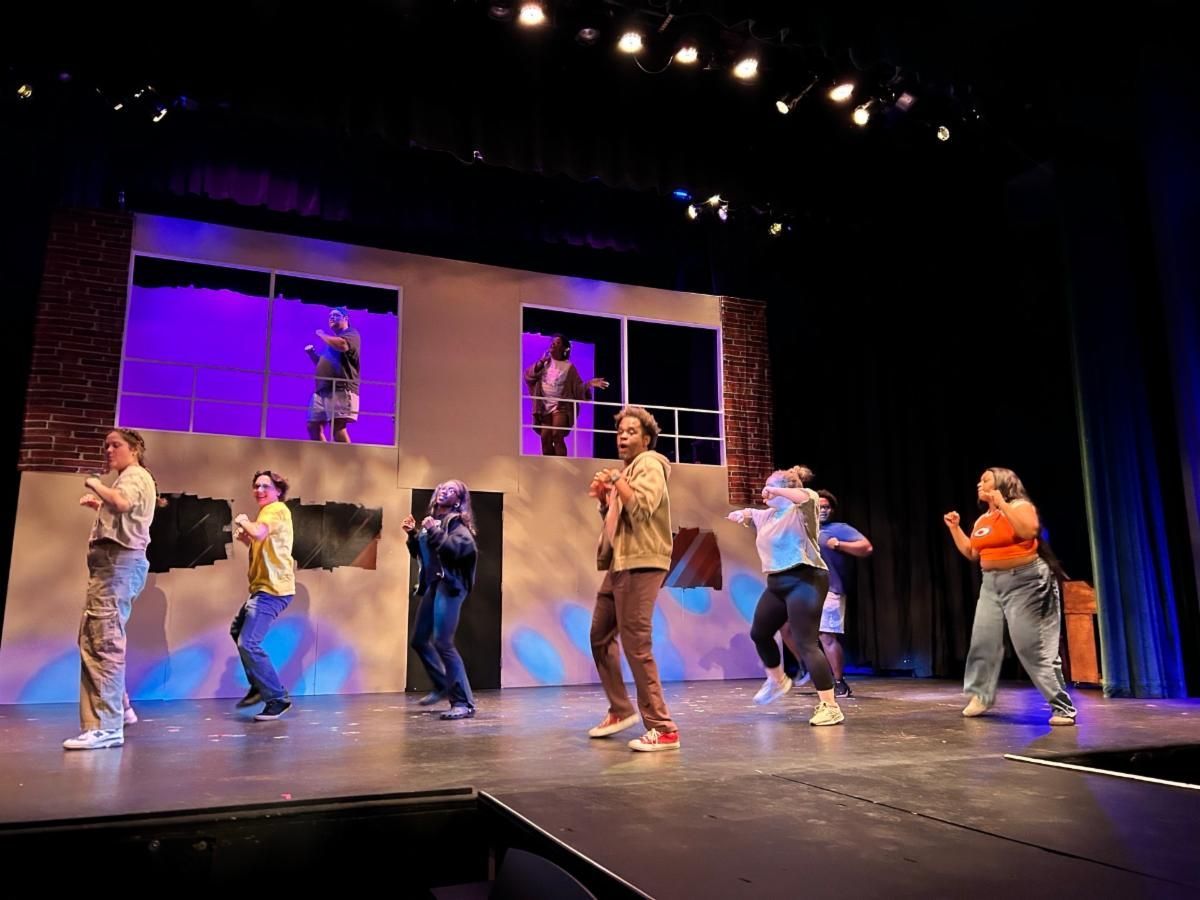 Dancers performing on a theater stage with a purple-lit set and brick backdrop