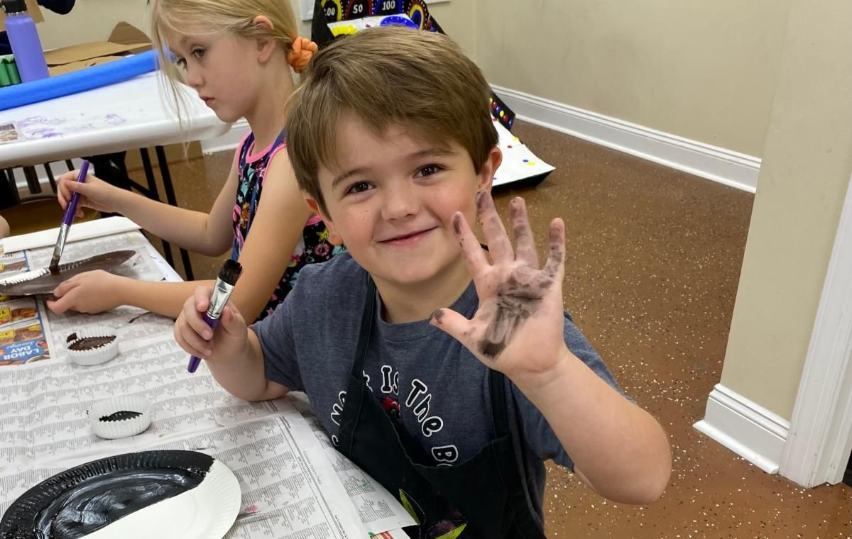 Child at an art table smiling and holding up a paint-covered hand, with another child painting in the background