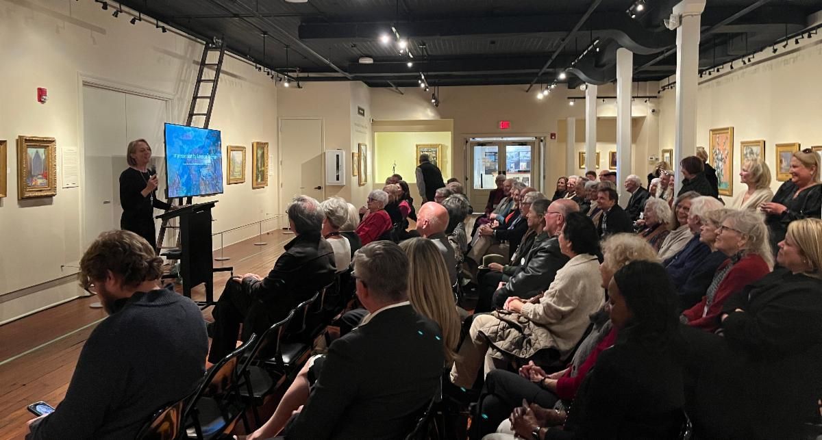 Audience seated in an art gallery listening to a speaker beside a blue-lit screen