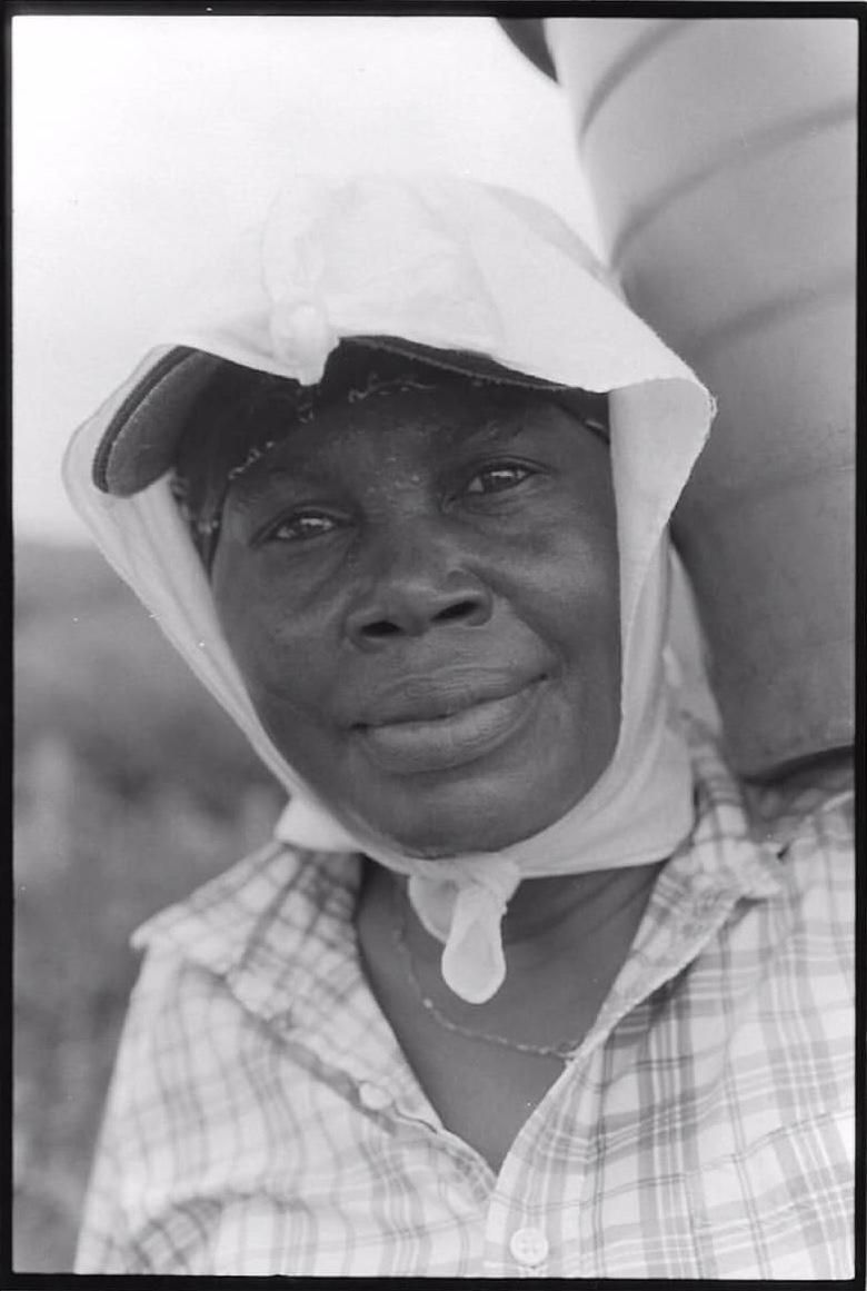 Person wearing a white head covering and checkered shirt, looking at the camera with a faint smile.