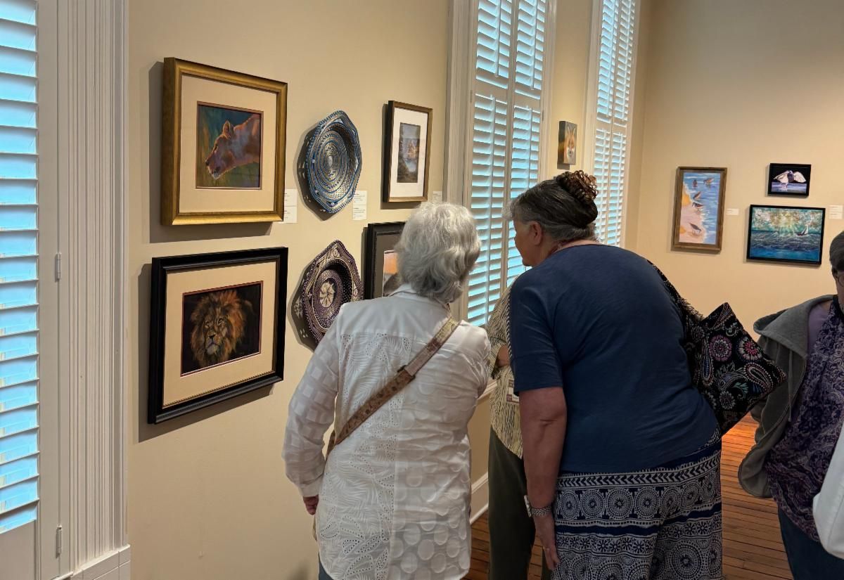 Three people viewing framed artwork in a gallery with white walls and shuttered windows.